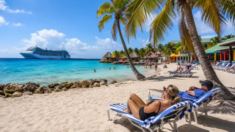 Cruise passengers relaxing on a beach near Cozumel cruise port with turquoise water, palm trees, beach chairs, and a cruise ship in the distance